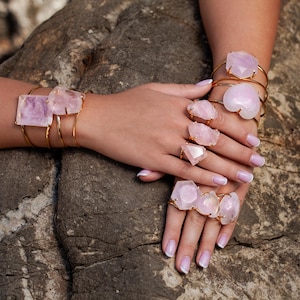 Puede incluir: Una colecci&oacute;n de joyas doradas con grandes piedras de cuarzo rosa en bruto. Las piezas incluyen pulseras y anillos, mostrando los tonos rosados naturales de los cristales. Las joyas se exhiben sobre una superficie de piedra.
