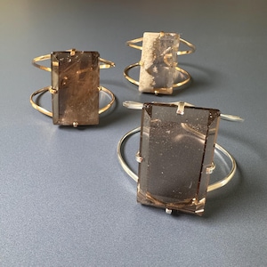 May include: Three cuff bracelets with rectangular, smoky quartz stones. The bracelets have gold and silver-toned metal bands. The quartz stones are a translucent brown color. The bracelets are displayed on a gray surface.