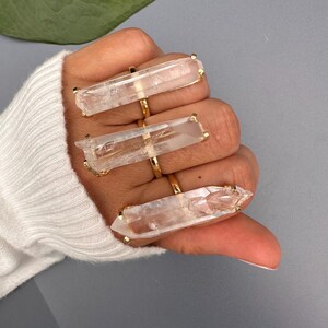 May include: Three gold rings, each with a large, clear quartz crystal. The crystals are rectangular with pointed ends. The rings are displayed on a hand wearing a white sweater, against a grey background.