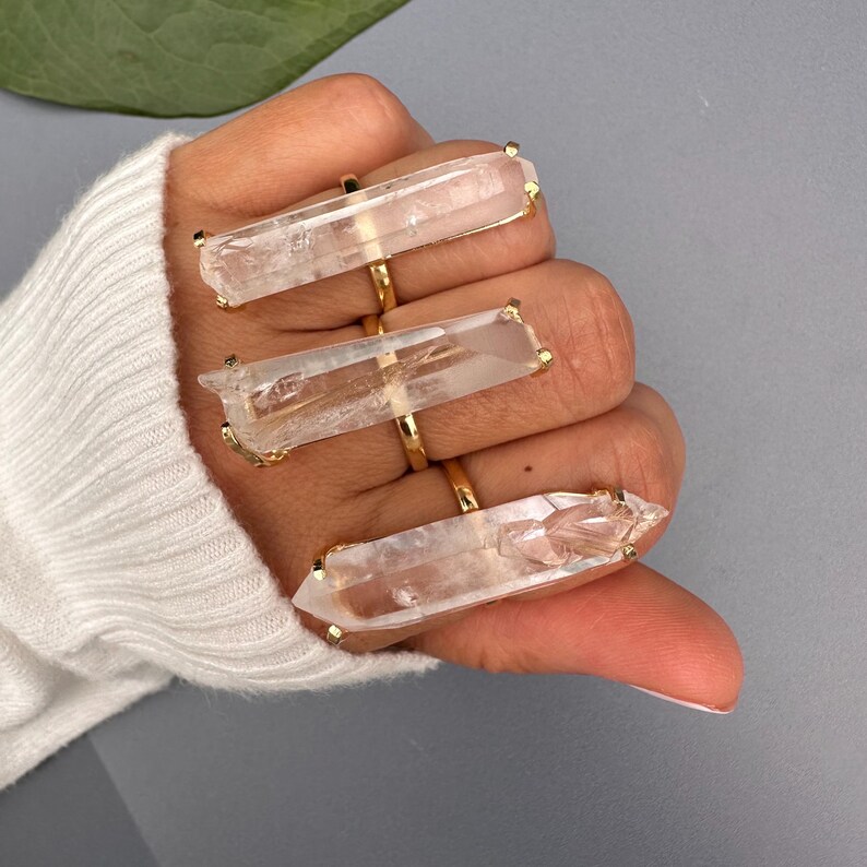 May include: Three gold rings, each with a large, clear quartz crystal. The crystals are rectangular with pointed ends. The rings are displayed on a hand wearing a white sweater, against a grey background.