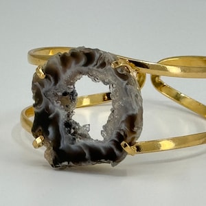 May include: A gold metal cuff bracelet with a large, dark gray agate stone featuring a natural crystal formation. The stone is set in the center of the bracelet and is surrounded by a gold metal band.
