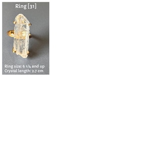 May include: A gold ring with a clear, rectangular crystal. The crystal measures approximately 2.7 cm in length. Ring size is 6 1/4 and up. The image includes the text "Ring [31]" and "Ring size: 6 1/4 and up Crystal length: 2.7 cm".