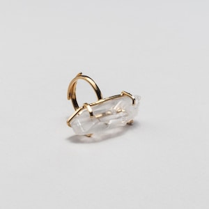 May include: A gold ring with a clear quartz crystal. The quartz is rectangular and set horizontally. The ring has a simple band and prongs to hold the crystal. The quartz is a natural, clear stone.