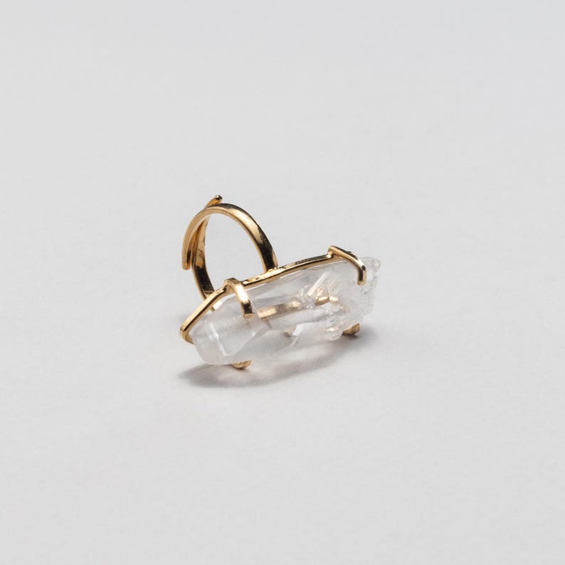 May include: A gold ring with a clear quartz crystal. The quartz is rectangular and set horizontally. The ring has a simple band and prongs to hold the crystal. The quartz is a natural, clear stone.