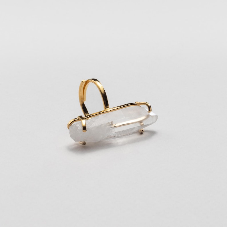 May include: A gold ring with a clear quartz crystal. The quartz is rectangular and set horizontally in a gold frame. The ring band is a simple, curved design.