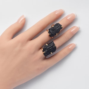 May include: Two rings featuring raw, vertically striated black tourmaline crystals set in gold-toned metal prongs. The stones have a rough, natural texture and are secured by four metal claws on each side, showcasing a dark, opaque appearance.