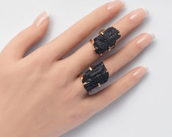 May include: Two rings featuring raw, vertically striated black tourmaline crystals set in gold-toned metal prongs. The stones have a rough, natural texture and are secured by four metal claws on each side, showcasing a dark, opaque appearance.