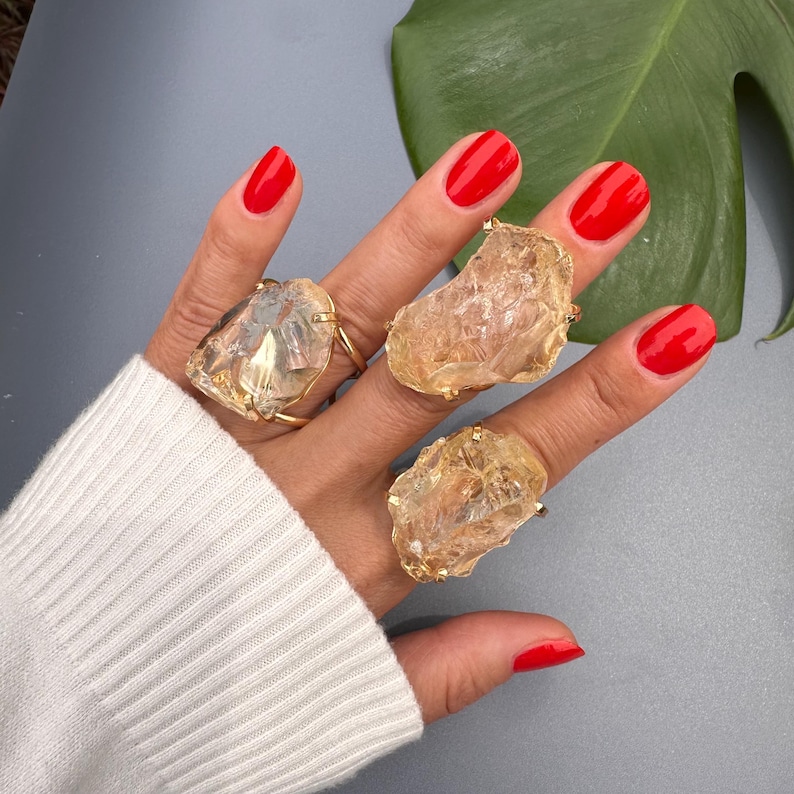 May include: Three gold rings each feature a large, irregularly shaped, translucent, amber-colored stone. The rings are displayed on a hand with red nail polish, against a gray background. The hand is wearing a white sweater.