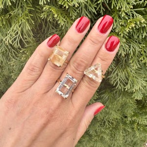 May include: Three gold rings with clear and golden quartz stones. The rings are on a hand with red nail polish.