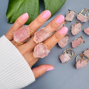 Puede incluir: Anillos de cuarzo rosa con bandas doradas. Los anillos presentan grandes piedras de cuarzo rosa de forma irregular. Varios anillos se muestran en una mano con esmalte de u&ntilde;as rosa, y otros est&aacute;n dispersos sobre una superficie gris.