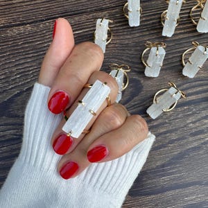 May include: A gold-toned ring featuring a long, rectangular, translucent white selenite crystal held by four metal prongs. Several identical rings are arranged nearby, each showcasing the same raw, fibrous crystal structure and metallic band.