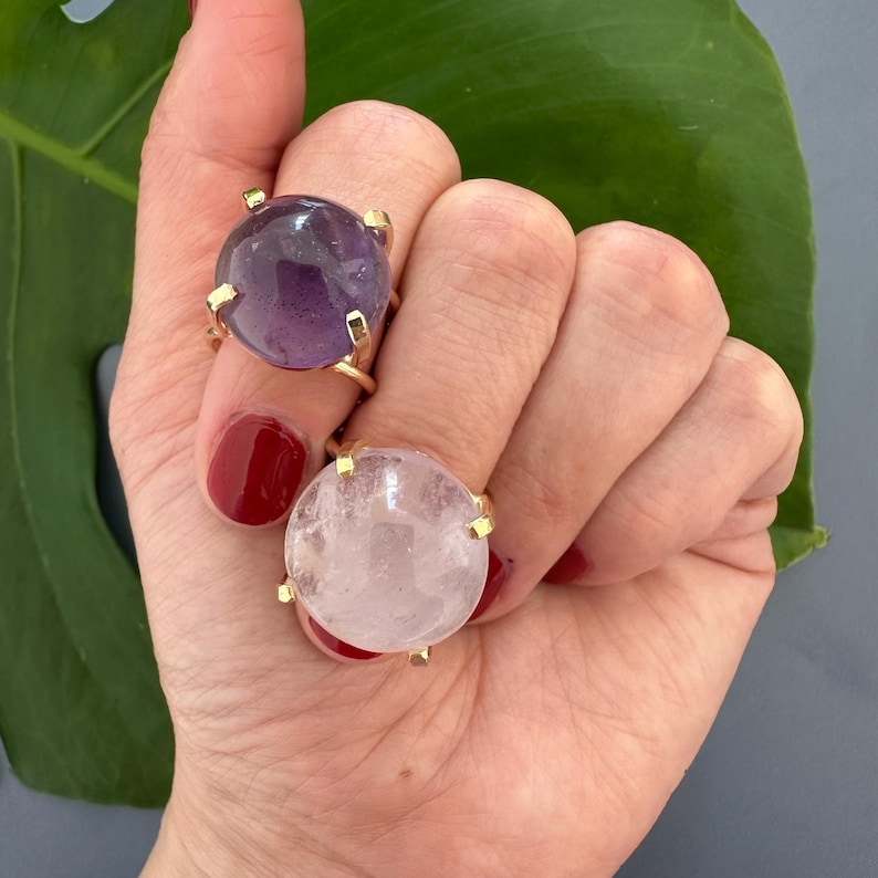 May include: Two gold rings with large round gemstone settings. The ring on the left has a purple amethyst stone. The ring on the right has a clear quartz stone.