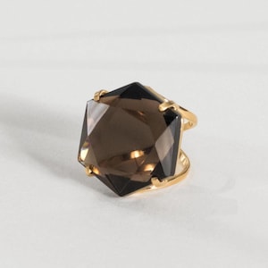 May include: A gold ring featuring a large, faceted, smoky quartz gemstone. The gemstone is a dark brown color and cut into a geometric shape. The ring has a simple, elegant design with a gold band.