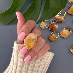 May include: A gold ring with a large, faceted, yellow citrine gemstone. The ring is shown on a hand wearing a cream-colored sweater.