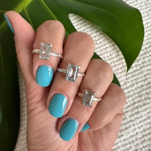 May include: Three silver rings with clear rectangular gemstones. The rings are worn on the fingers of a hand with turquoise nail polish.