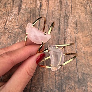 May include: Two gold metal bracelets with a crescent moon shaped rose quartz stone. The bracelets are stacked on top of each other.