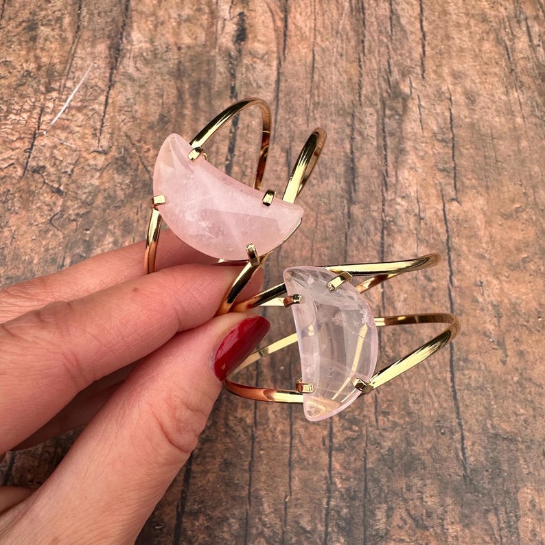 May include: Two gold metal bracelets with a crescent moon shaped rose quartz stone. The bracelets are stacked on top of each other.