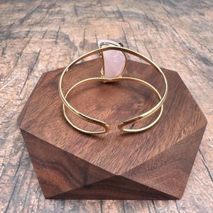 May include: A gold bangle bracelet with a large, light pink, semi-circular stone set into the front. The bracelet is open and rests on a wooden surface.