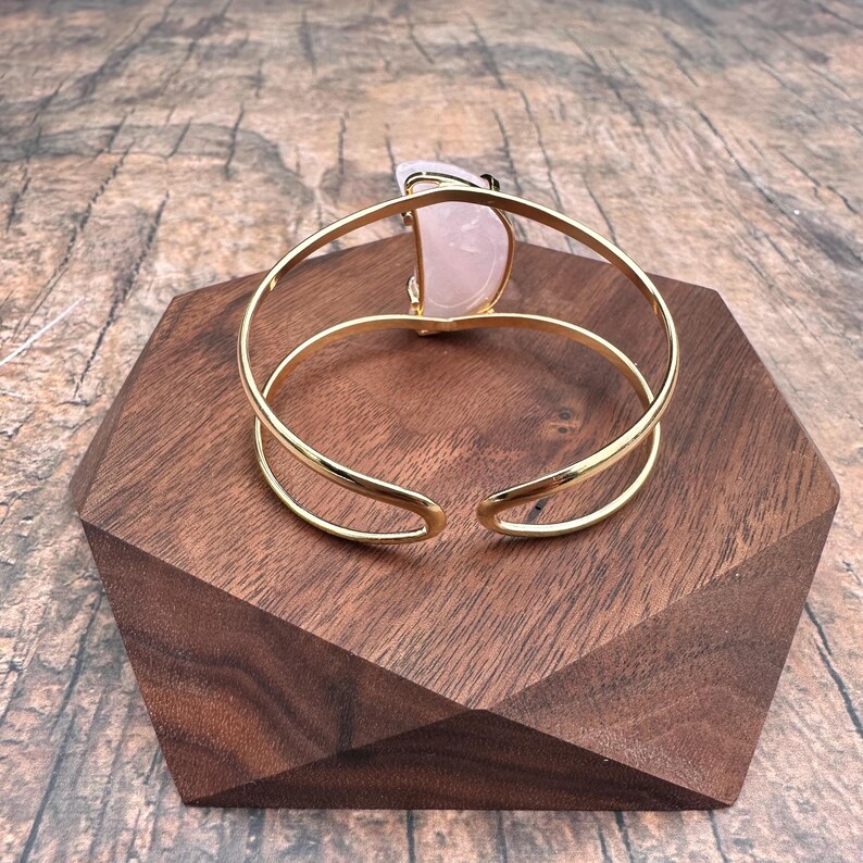 May include: A gold bangle bracelet with a large, light pink, semi-circular stone set into the front. The bracelet is open and rests on a wooden surface.