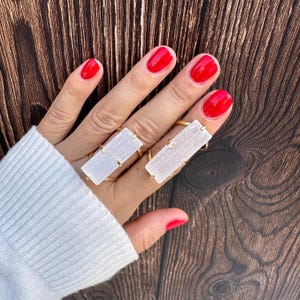 May include: Two gold-toned rings featuring rectangular, translucent white selenite crystal bars. Each ring has a thin metal band that secures the elongated, fibrous-textured stone horizontally across the finger, creating a distinct, layered appearance.