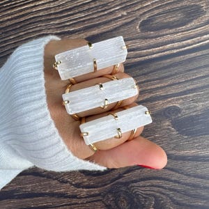 May include: Three rings featuring rectangular, translucent white selenite bars set horizontally in gold-toned metal prongs. Each ring has a smooth, polished band and is worn on three fingers, showcasing the fibrous, crystalline texture of the stone.