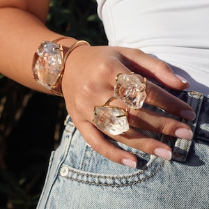 May include: A gold-toned bracelet and two rings featuring large, irregularly shaped, clear crystal stones. The jewelry is set against a background of denim jeans and a white top, creating a contrast of textures and colors.