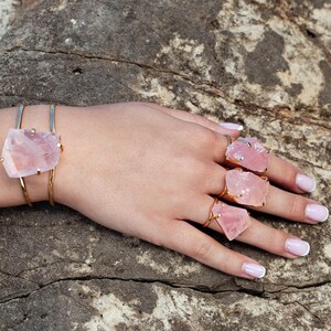 Puede incluir: Una pulsera y anillos dorados con grandes piedras de cuarzo rosa en bruto. La pulsera tiene una sencilla banda dorada, mientras que los anillos tienen engastes dorados. Las joyas se exhiben en una mano con u&ntilde;as cuidadas, sobre un fondo de piedra texturizada.