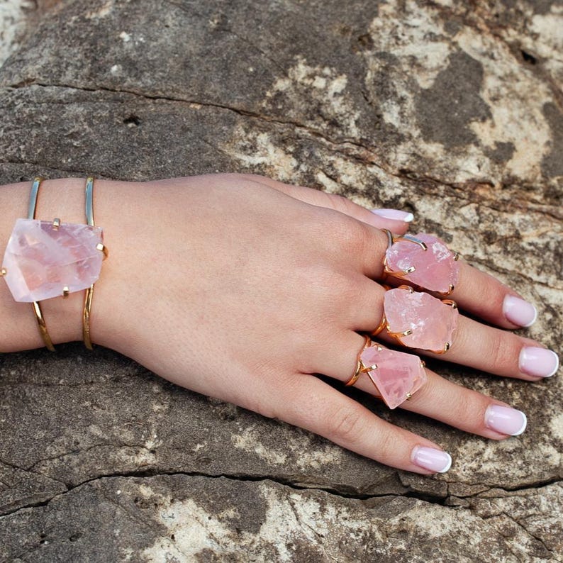 Puede incluir: Una pulsera y anillos dorados con grandes piedras de cuarzo rosa en bruto. La pulsera tiene una sencilla banda dorada, mientras que los anillos tienen engastes dorados. Las joyas se exhiben en una mano con u&ntilde;as cuidadas, sobre un fondo de piedra texturizada.