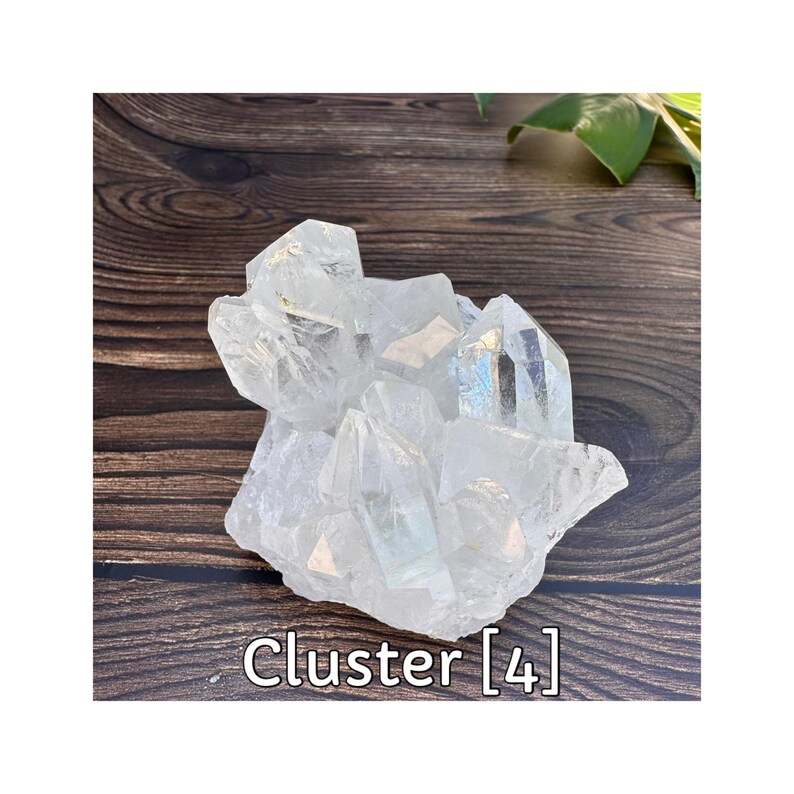 May include: A clear quartz crystal cluster with multiple points. The crystals are translucent with some areas showing iridescent colors. The cluster is sitting on a dark brown wooden surface. The text "Cluster [4]" is at the bottom.