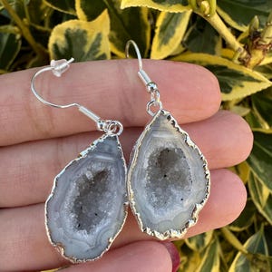 May include: A pair of silver earrings with a teardrop-shaped agate stone. The agate is a natural crystal with a white and gray color and a hollow center.