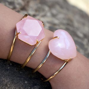 May include: Two rose quartz bracelets. One bracelet features a faceted, geometric pink stone. The other bracelet has a heart-shaped pink stone. Both stones are set in gold-colored metal and are worn on a wrist.