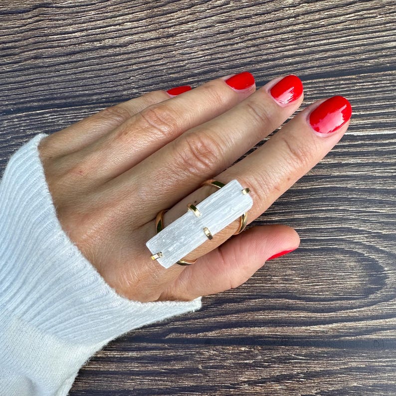 May include: A gold-toned ring featuring a rectangular, translucent white selenite crystal bar set horizontally across the finger. The crystal is secured by four small gold-toned metal prongs at the corners, creating a bold, geometric statement piece.