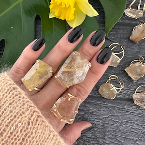 May include: A set of five gold rings featuring clear quartz crystals with a golden sheen. The rings are adjustable and feature a unique, faceted design.