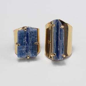 May include: Two gold-toned rings featuring raw, rectangular blue kyanite gemstones set in metal prongs. The ring on the left has a wider, shorter stone, while the ring on the right features a longer, narrower blue crystal set in a smooth band.