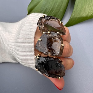 May include: Three rings with large, rough-cut, smoky quartz stones set in gold-colored metal. The rings are stacked on a finger, showcasing the dark brown, translucent crystals. The setting has a simple, geometric design.