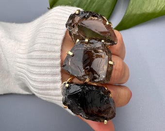 May include: Three rings featuring large, rough-cut, smoky quartz stones set in gold-coloured metal. The rings are stacked on a finger, showcasing the dark brown, translucent crystals. The setting has a simple, geometric design.