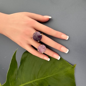 May include: Two gold rings with purple and light purple round gemstones. The rings are on a hand with white nail polish.