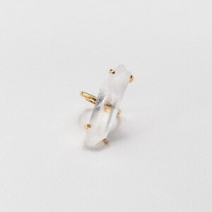 May include: A gold ring with a clear, elongated crystal. The crystal is mounted vertically on the ring, with gold accents securing it. The ring is set against a plain white background, highlighting the jewelry's design and materials.