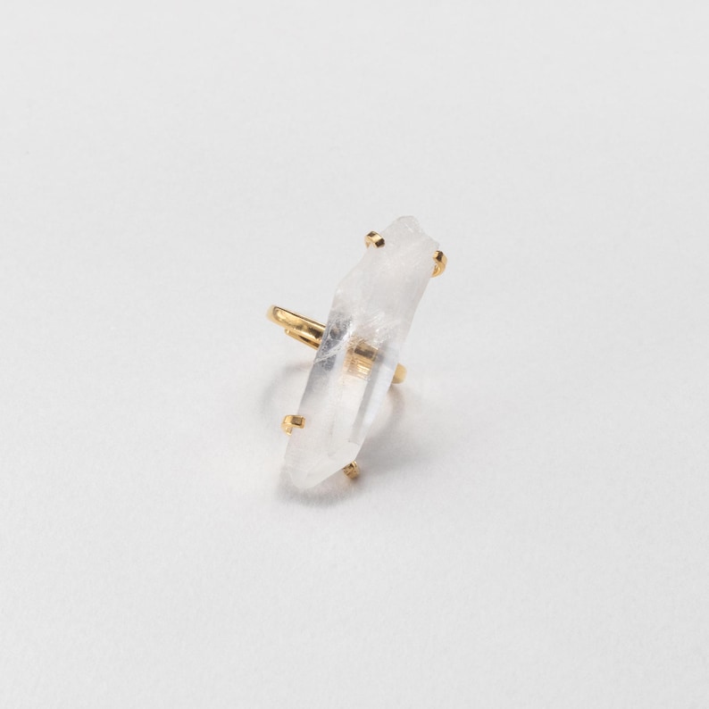 May include: A gold ring with a clear, elongated crystal. The crystal is mounted vertically on the ring, with gold accents securing it. The ring is set against a plain white background, highlighting the jewelry's design and materials.