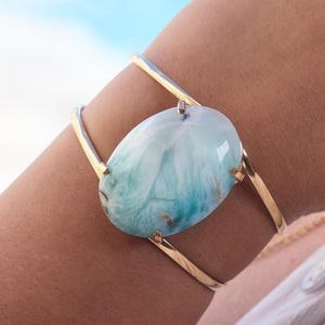 May include: A gold-toned cuff bracelet featuring a large, oval-shaped, polished light blue and white stone with natural marbling patterns. The stone is secured by four small metal prongs, and the band consists of two thin, smooth, open-ended metal wires.