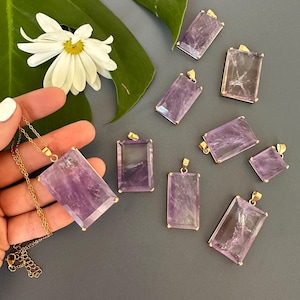 May include: A collection of rectangular amethyst pendants with gold-colored settings. One pendant is on a gold chain. The pendants vary in size and are displayed on a gray surface, with a white daisy and green leaf in the background.