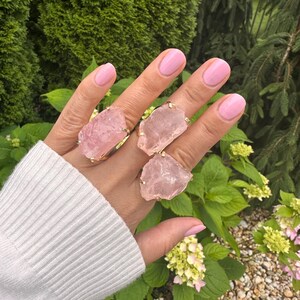 Puede incluir: Cuatro anillos de cuarzo rosa con bandas doradas se muestran en una mano. Los anillos presentan piedras grandes de forma irregular en varios tonos de rosa. El fondo incluye follaje verde y un su&eacute;ter blanco.