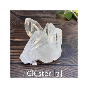 May include: A clear quartz crystal cluster with multiple points. The crystal is transparent with visible internal structures. The base is a rough, opaque white. The text "Cluster [3]" is at the bottom.