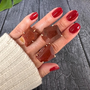 May include: Three gemstone rings in shades of red and brown are displayed. The rings feature large, faceted stones set in gold-colored metal. The hand wearing the rings has red nail polish and is wearing a cream-colored sweater.