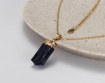 May include: A gold chain necklace featuring a raw, vertical black tourmaline crystal pendant. The top of the crystal is encased in a textured gold cap, and the necklace includes a delicate gold link chain with a small oval tag near the clasp.