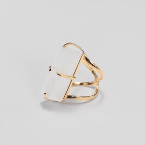 May include: A ring featuring a rectangular, translucent white selenite crystal set in a polished gold-tone metal band. The crystal is held in place by four thin metal prongs, and the band consists of two delicate, curved gold-tone wires.