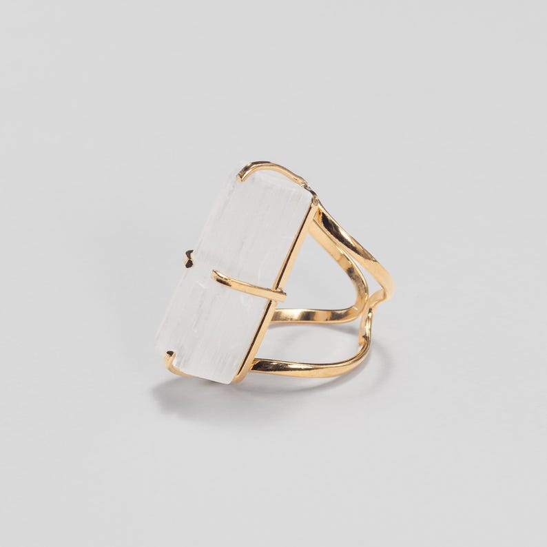 May include: A ring featuring a rectangular, translucent white selenite crystal set in a polished gold-tone metal band. The crystal is held in place by four thin metal prongs, and the band consists of two delicate, curved gold-tone wires.