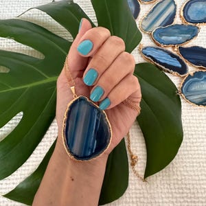 May include: A gold chain necklace with a large blue agate pendant. The agate is a natural stone with white and blue stripes. The pendant is in the shape of a teardrop.