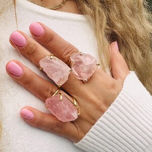 Puede incluir: Tres anillos de cuarzo rosa con bandas doradas se muestran en una mano. Los anillos presentan piedras de color rosa claro de forma irregular. La persona lleva un su&eacute;ter blanco y tiene esmalte de u&ntilde;as rosa.