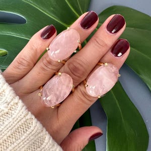 May include: Three gold rings with oval shaped rose quartz stones. The rings are worn on a hand with dark red nail polish.
